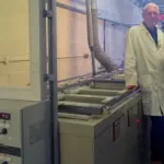 Photograph of three men, two in long white lab coats and one wearing a long blue coat stand next to a grey PCB plating machine, the tops of four tanks just visible.